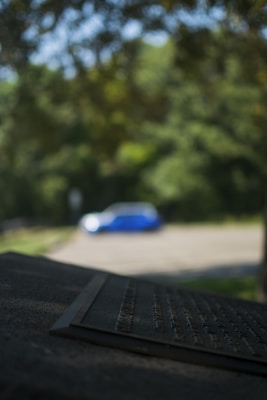 A blurred image displaying a metallic plaque in the foreground with engraved text, partially in focus. In the background, there is a blue and white object, possibly a car, situated in a parking area surrounded by lush greenery and trees.