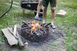 A person wearing flip-flops is crouched near a fire pit, using a canister to help ignite a small fire made of gathered twigs and branches. A wheelbarrow filled with additional wood and sticks is visible in the background on a grassy lawn. Nearby lies a pile of extra wood and an open cardboard box.