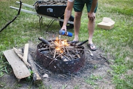 A person wearing flip-flops is crouched near a fire pit, using a canister to help ignite a small fire made of gathered twigs and branches. A wheelbarrow filled with additional wood and sticks is visible in the background on a grassy lawn. Nearby lies a pile of extra wood and an open cardboard box.