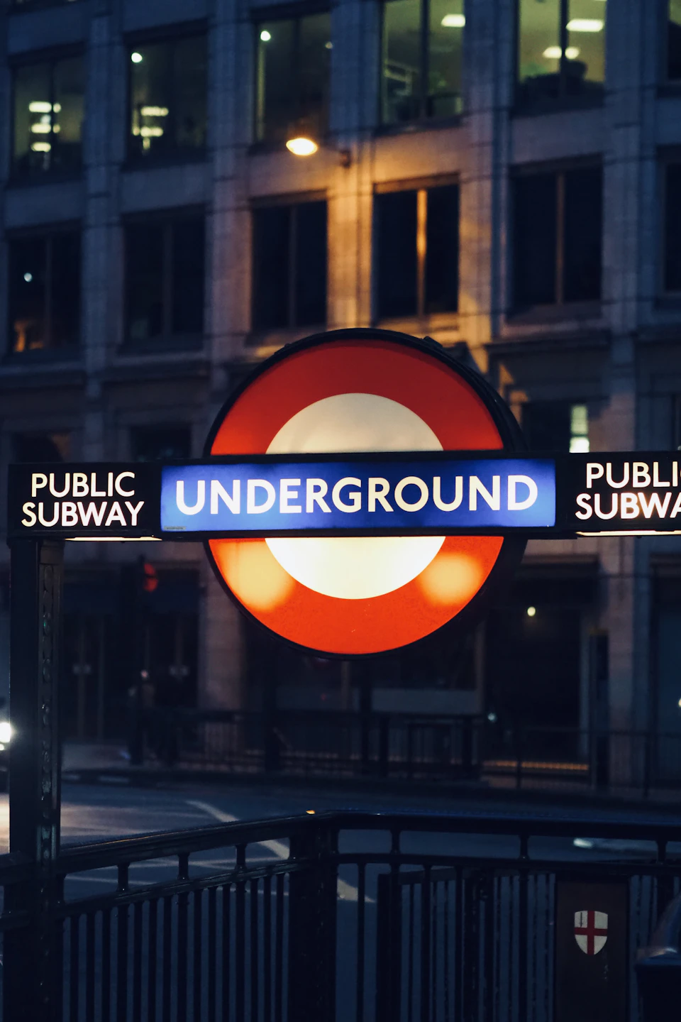 The image shows a street scene at night, with a prominent Underground sign mounted on a black pole. The sign is illuminated by a red and white light, making it easily visible against the dark background. Below the sign, there are two signs indicating the Underground's location - one for "Public Subway" and another for "Underground". In the foreground, there is a metal railing that adds depth to the scene.
