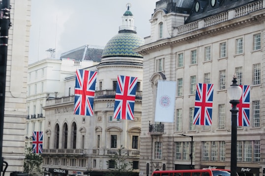 A city street scene with several British flags hanging from buildings. The architecture is classic with ornate details and a prominent domed building. A red double-decker bus is partially visible at the bottom, indicating a typical urban setting.