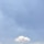 A single, fluffy white cloud is floating in a vast, clear blue sky. Below the cloud, a tall communication tower is visible.