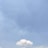 A single, fluffy white cloud is floating in a vast, clear blue sky. Below the cloud, a tall communication tower is visible.