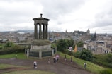 A large classical monument with columns stands prominently in a park setting overlooking a cityscape. People are walking around near the monument. The city is characterized by historic and traditional architecture, with numerous spires and stone buildings. The sky is overcast, creating a calm and subdued atmosphere.