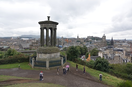 A large classical monument with columns stands prominently in a park setting overlooking a cityscape. People are walking around near the monument. The city is characterized by historic and traditional architecture, with numerous spires and stone buildings. The sky is overcast, creating a calm and subdued atmosphere.