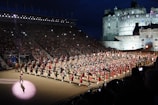 A large organized parade featuring numerous participants dressed in traditional military uniforms with kilts, accompanied by a variety of instruments. The event takes place in an outdoor venue with grandstands full of spectators. A historic castle serves as the backdrop under a dark evening sky, illuminated by lights.