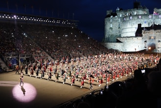 A large organized parade featuring numerous participants dressed in traditional military uniforms with kilts, accompanied by a variety of instruments. The event takes place in an outdoor venue with grandstands full of spectators. A historic castle serves as the backdrop under a dark evening sky, illuminated by lights.