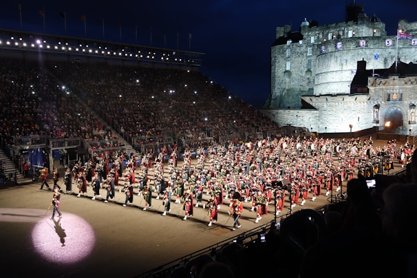 A large organized parade featuring numerous participants dressed in traditional military uniforms with kilts, accompanied by a variety of instruments. The event takes place in an outdoor venue with grandstands full of spectators. A historic castle serves as the backdrop under a dark evening sky, illuminated by lights.
