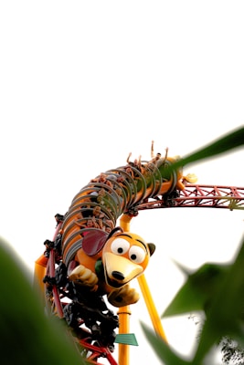 A colorful roller coaster is depicted with cars designed in the theme of a cartoon character styled as a stretching dog. Passengers have their arms raised, indicating excitement and thrill, as the ride takes a sharp curve. The image is framed by green foliage, adding an element of nature to the scene.