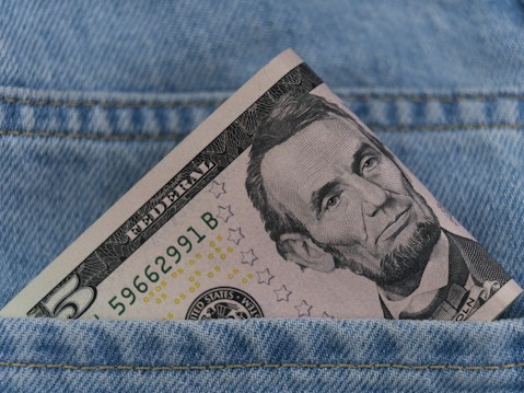 A five-dollar bill is partially visible, tucked into the front pocket of blue denim jeans. The bill is prominently featuring a portrait of a historical figure.
