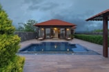 A luxurious villa with a red-tiled roof is set against a backdrop of dark, cloudy skies. There is a clear blue swimming pool in the patio area surrounded by a wooden deck. The lush greenery and landscaped plants border the patio, adding to the tranquil atmosphere.