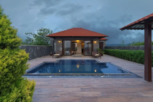 A luxurious villa with a red-tiled roof is set against a backdrop of dark, cloudy skies. There is a clear blue swimming pool in the patio area surrounded by a wooden deck. The lush greenery and landscaped plants border the patio, adding to the tranquil atmosphere.