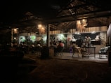 A dimly lit restaurant or café with a rustic interior. There are people sitting at tables under a wooden canopy with hanging lamps. The back wall features several large plants, adding a touch of greenery. Three people are standing near a counter, possibly preparing or ordering food or drinks.