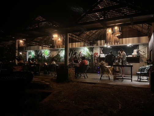 A dimly lit restaurant or café with a rustic interior. There are people sitting at tables under a wooden canopy with hanging lamps. The back wall features several large plants, adding a touch of greenery. Three people are standing near a counter, possibly preparing or ordering food or drinks.
