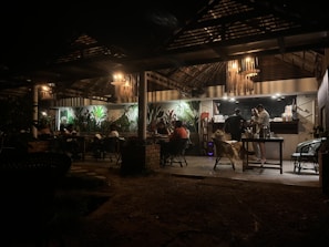 A dimly lit restaurant or café with a rustic interior. There are people sitting at tables under a wooden canopy with hanging lamps. The back wall features several large plants, adding a touch of greenery. Three people are standing near a counter, possibly preparing or ordering food or drinks.