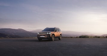 A modern SUV is parked on an open road with a mountainous landscape in the background. The vehicle is highlighted against the soft, muted colors of the sunset sky, contributing to a serene and adventurous atmosphere.
