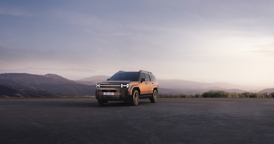 A modern SUV is parked on an open road with a mountainous landscape in the background. The vehicle is highlighted against the soft, muted colors of the sunset sky, contributing to a serene and adventurous atmosphere.