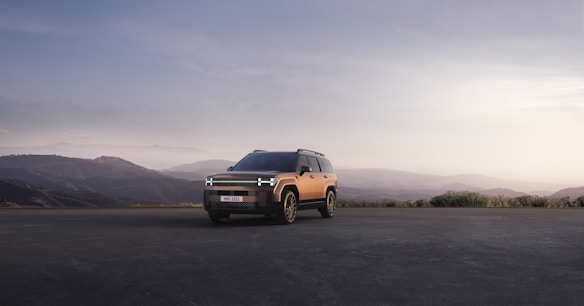 A modern SUV is parked on an open road with a mountainous landscape in the background. The vehicle is highlighted against the soft, muted colors of the sunset sky, contributing to a serene and adventurous atmosphere.
