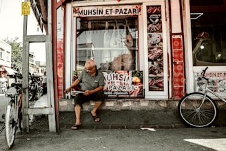A man sits outside a butcher shop, reading or handling something in front of him. The shop displays various images of meat products on its windows, and two bicycles are parked nearby. The scene is set on a street with other buildings visible in the background.