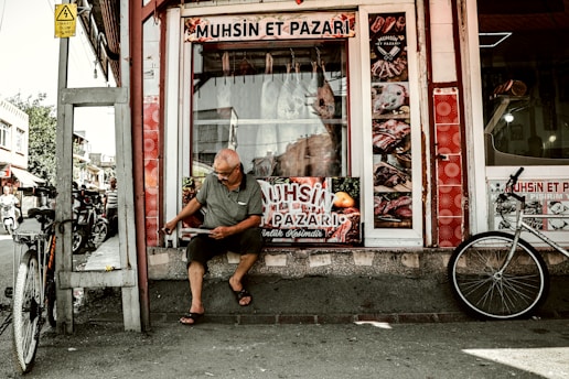 A man sits outside a butcher shop, reading or handling something in front of him. The shop displays various images of meat products on its windows, and two bicycles are parked nearby. The scene is set on a street with other buildings visible in the background.
