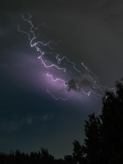 A vivid lightning strike illuminates the night sky, casting light through a backdrop of dark clouds. Silhouetted trees appear at the bottom, creating a contrast between the illuminated sky and the darkness of the landscape.