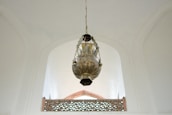 A large, intricately designed brass lantern hangs from the ceiling of a spacious and well-lit room with high, arched ceilings. The lantern features detailed patterns and artistic craftsmanship. Below, there is an ornate railing with a geometric pattern that aligns with the architectural style of the room.