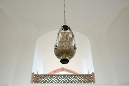 A large, intricately designed brass lantern hangs from the ceiling of a spacious and well-lit room with high, arched ceilings. The lantern features detailed patterns and artistic craftsmanship. Below, there is an ornate railing with a geometric pattern that aligns with the architectural style of the room.