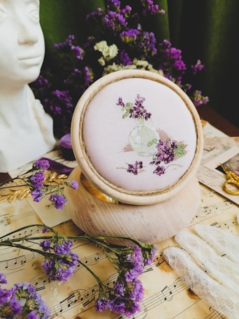 A close-up of an embroidered fabric with purple flowers displayed in a wooden hoop. The setup includes a bouquet of purple flowers scattered on a vintage sheet of music, a lace glove, a bust statue, golden scissors, and a spool of thread in the background.