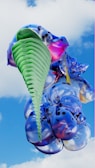 An abstract 3D sculpture rendered against a backdrop of a blue sky with clouds. The sculpture features a combination of vibrant colors including green, blue, purple, and pink. Its form resembles a cluster of glossy, smooth, and inflated shapes intertwined with a spiral, ribbed structure.
