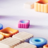 A collection of pastel-colored, geometrically shaped objects placed on a white grid surface. The objects include various shapes like squares, circles, and rectangles, made from a glossy material with a soft, smooth appearance. The central focus is a pink square block with rounded edges and a subtle blue gradient in the center.