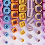 An arrangement of geometrically shaped, colorful 3D objects featuring vibrant colors like pink, blue, orange, and yellow. The objects include rings, squares, and circular shapes with textures ranging from smooth to striped. The composition creates a visually appealing pattern set against a grid-like background.