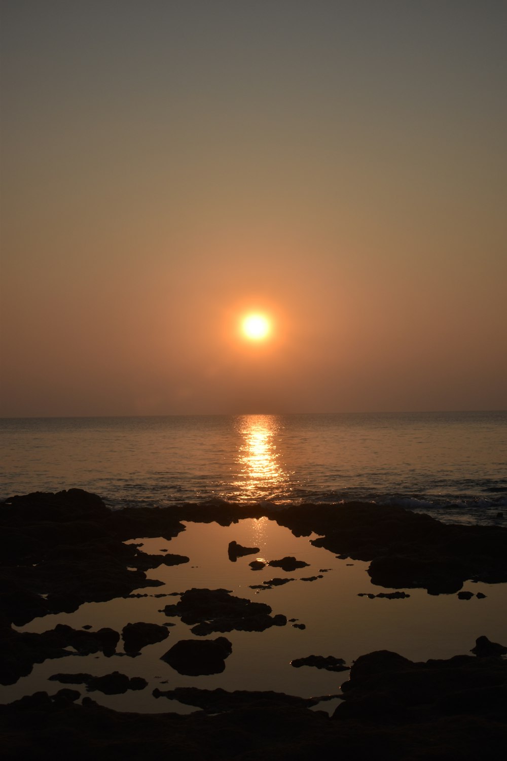 the sun is setting over the ocean with rocks in the foreground