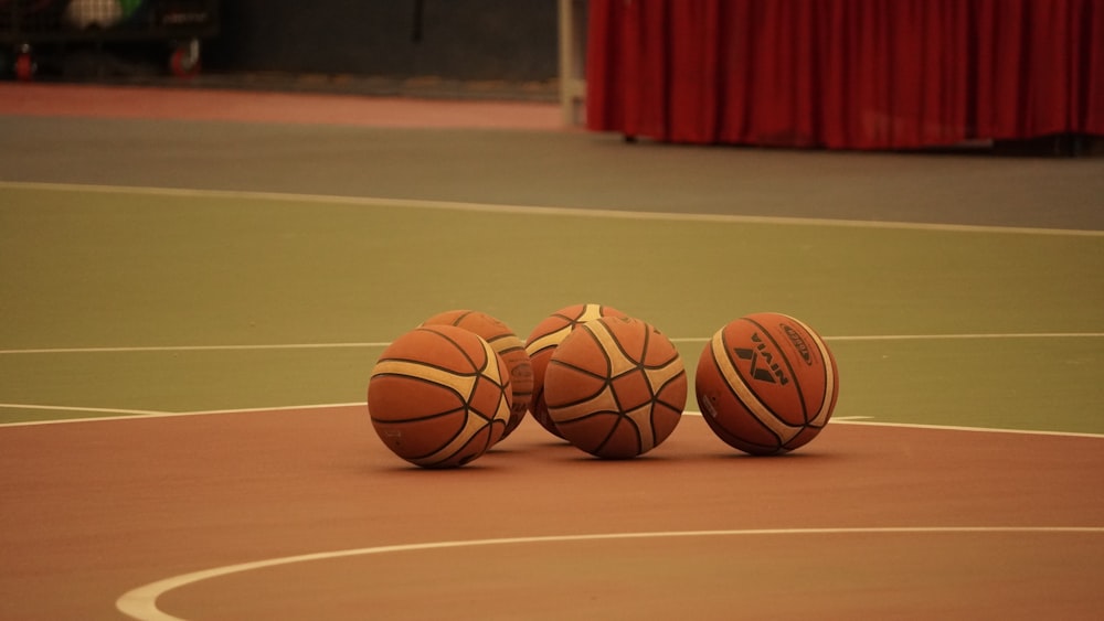 três bolas de basquete sentadas em uma quadra de basquete com uma cortina vermelha ao fundo