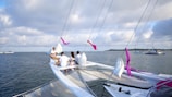 A group of people is sitting on the bow of a trimaran sailboat on a large body of water, with other sailboats visible in the distance. The sky is partly cloudy, creating a calm and serene atmosphere. The sailboat has purple ribbons tied to the rigging.