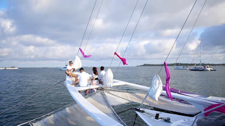 A group of people is sitting on the bow of a trimaran sailboat on a large body of water, with other sailboats visible in the distance. The sky is partly cloudy, creating a calm and serene atmosphere. The sailboat has purple ribbons tied to the rigging.