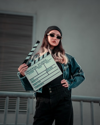 A stylish individual wearing sunglasses and a black beanie holds a film clapperboard. Their attire includes a denim jacket paired with dark pants. The background is a muted color, suggesting an urban setting possibly near a production site.
