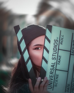 A person partially obscured by a large clapperboard, with only one eye visible. The clapperboard has text that reads 'Universal Studios' along with other typical film slate labels like scene, take, and roll. The background is blurred, suggesting an outdoor setting.