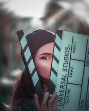A person partially obscured by a large clapperboard, with only one eye visible. The clapperboard has text that reads 'Universal Studios' along with other typical film slate labels like scene, take, and roll. The background is blurred, suggesting an outdoor setting.