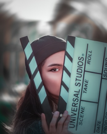 A person partially obscured by a large clapperboard, with only one eye visible. The clapperboard has text that reads 'Universal Studios' along with other typical film slate labels like scene, take, and roll. The background is blurred, suggesting an outdoor setting.