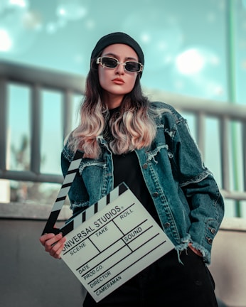 A person wearing a denim jacket, dark sunglasses, and a black beanie is holding a clapperboard used in film production. The background is blurred with a cool tone, featuring a railing.