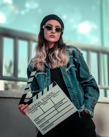 A person wearing a denim jacket, dark sunglasses, and a black beanie is holding a clapperboard used in film production. The background is blurred with a cool tone, featuring a railing.