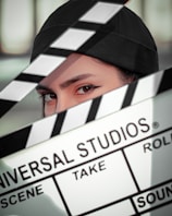 A person wearing a black cap is partially obscured by a film clapperboard. The clapperboard is prominently displaying the text 'Universal Studios' along with scene and take details. The person's eyes are sharply focused.