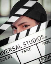 A person wearing a black cap is partially obscured by a film clapperboard. The clapperboard is prominently displaying the text 'Universal Studios' along with scene and take details. The person's eyes are sharply focused.