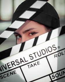 A person wearing a black cap is partially obscured by a film clapperboard. The clapperboard is prominently displaying the text 'Universal Studios' along with scene and take details. The person's eyes are sharply focused.