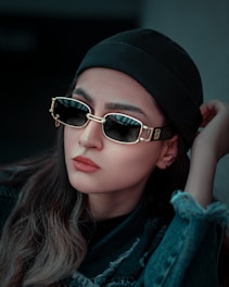 A person wearing stylish sunglasses with a gold frame and a black beanie. The individual has long hair and is dressed in a dark, rugged jacket, looking away from the camera with a thoughtful expression.