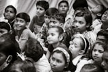 A group of children gather closely together, attentively observing something. Their expressions vary from curiosity to wonder. The scene is in black and white, creating a timeless feel.