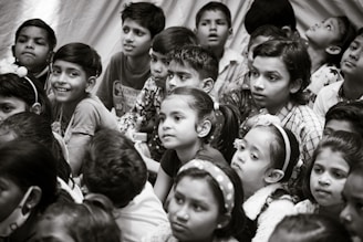 A group of children gather closely together, attentively observing something. Their expressions vary from curiosity to wonder. The scene is in black and white, creating a timeless feel.