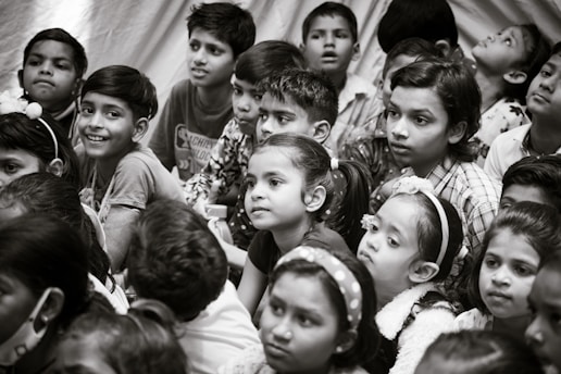 A group of children gather closely together, attentively observing something. Their expressions vary from curiosity to wonder. The scene is in black and white, creating a timeless feel.