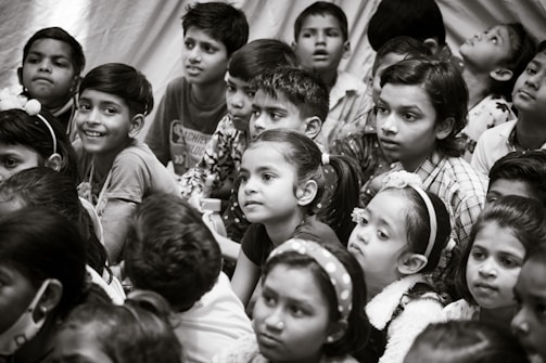 A group of children gather closely together, attentively observing something. Their expressions vary from curiosity to wonder. The scene is in black and white, creating a timeless feel.