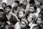 A group of children gather closely together, attentively observing something. Their expressions vary from curiosity to wonder. The scene is in black and white, creating a timeless feel.